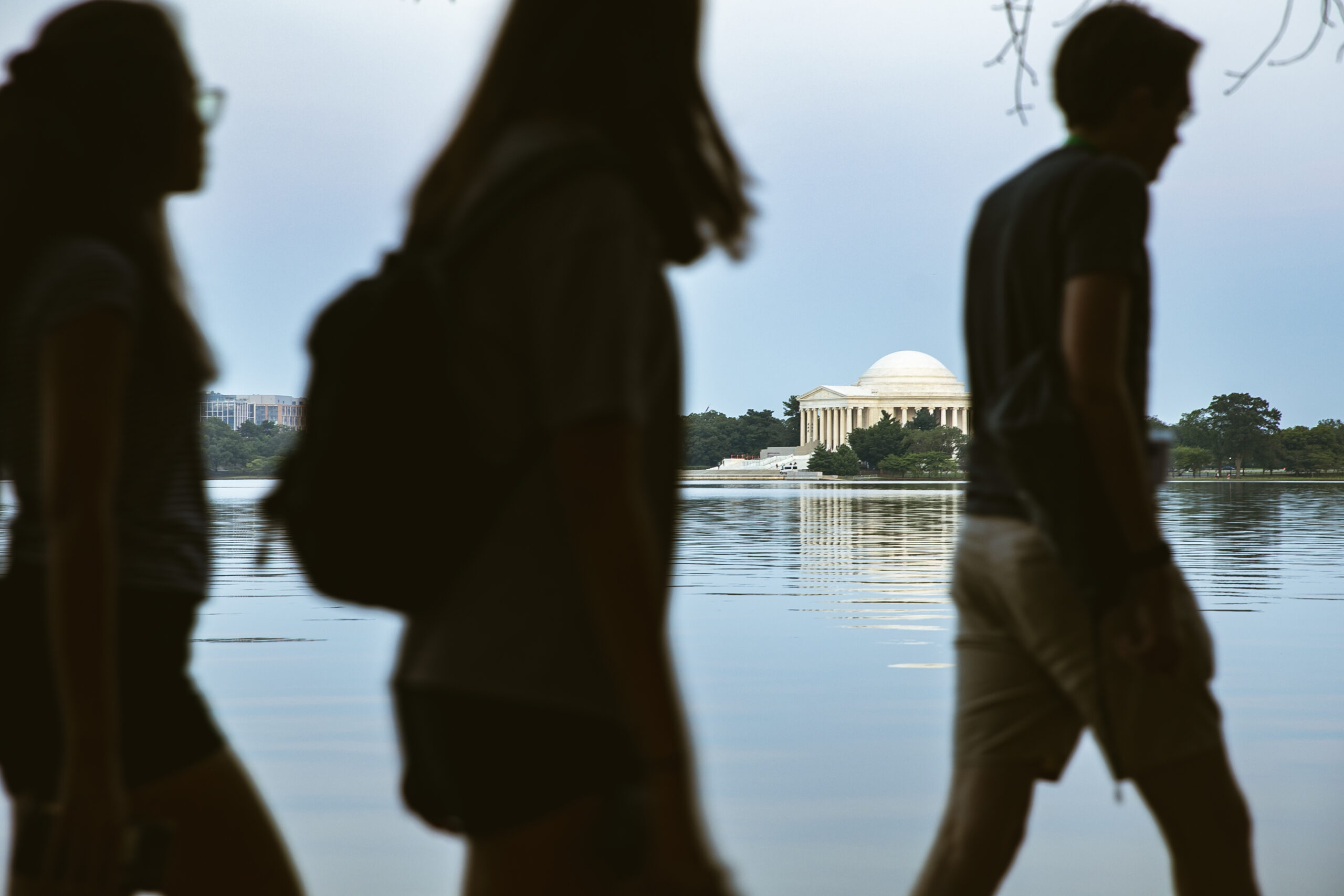 Three people with blurred and shadowed silhouettes appear in the foreground of the photo. Behind them, the Jefferson Memorial in D.C. can be seen in focus. The photo takes place on a summer day, and the memorial is reflect on the blue water of the tidal basin in Washington, D.C.