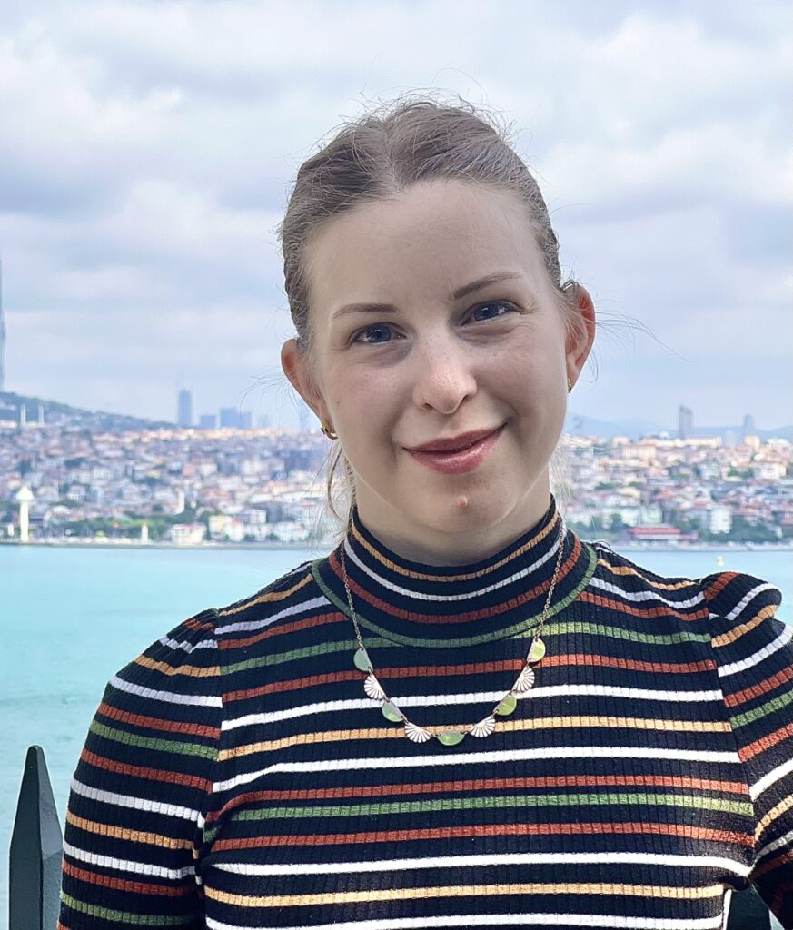 Audrey, a white woman with blonde hair, is pictured with a view of the Bosphorus and the Istanbul shoreline in the background. She wears a black turtle neck with horizontal stripes in alternating green, red, white, and yellow.