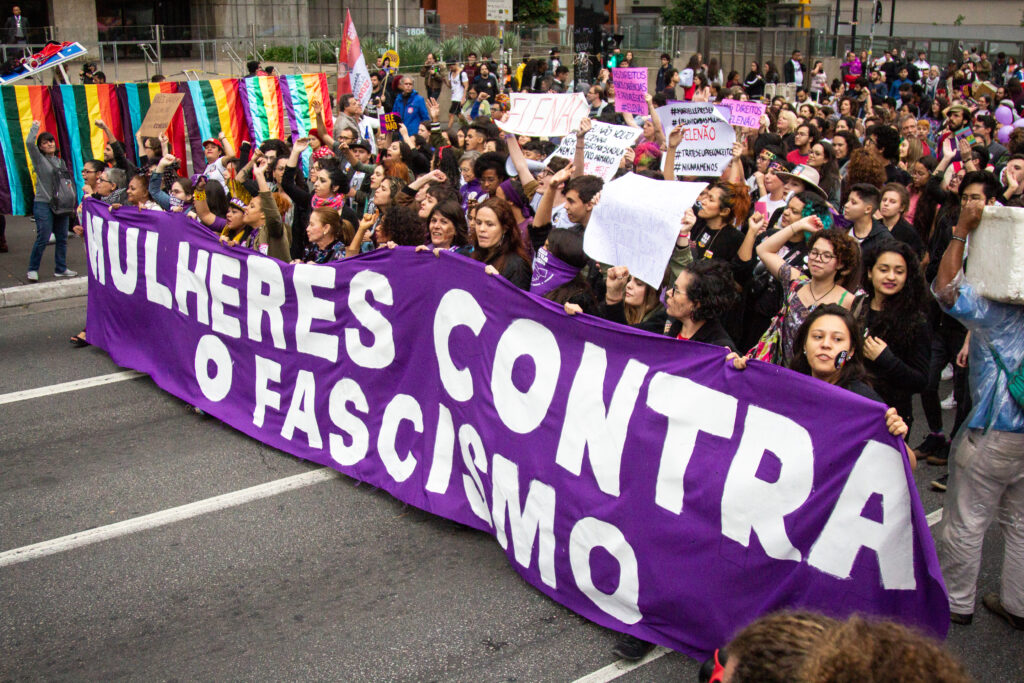 A large group of women and men holding a purple banner that says "Mulheres Contra O Fascismo" (Women Against Fascism) marches down a street as part of the #EleNao protests against Bolsonaro.