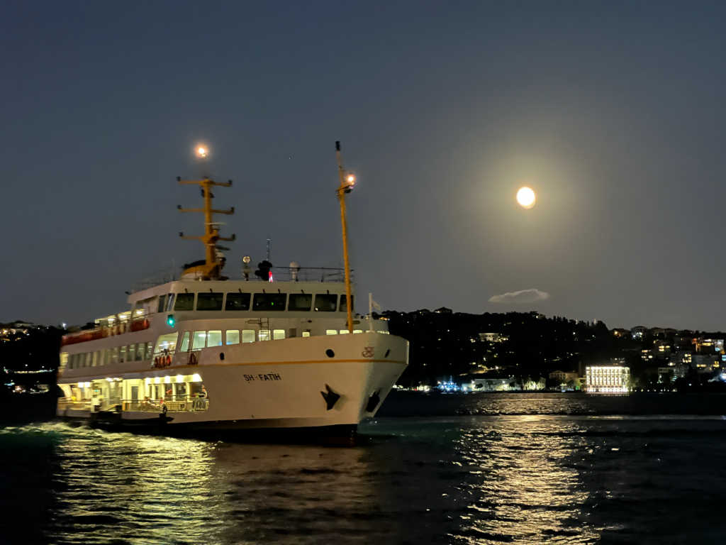 On a clear summer night with a brilliant full moon, a classic Istanbul ferry traverses the Bosphorus, approaching port. The lights of the ferry, the lights of Istanbul's Asian shore, and the light of the moon dance on the dark waters of the Bosphorus.