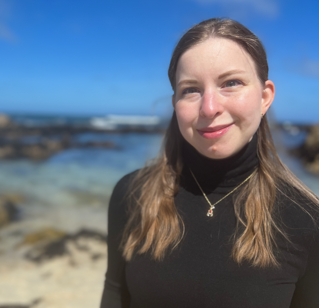 Portrait-style photo of Audrey Williams, a white woman with long dirty-blonde hair. She is wearing a black turtle-neck and a small gold and red necklace. She is pictured in front of the ocean at Pacific Grove, California.
