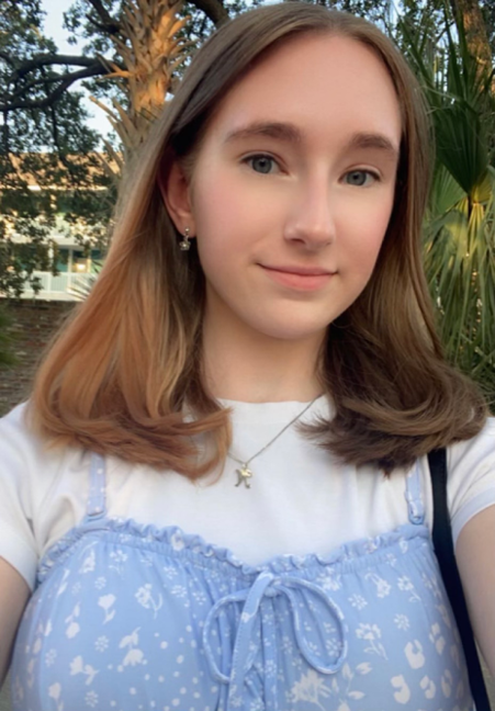Selfie-style photo of Nicole Struzinsky, a white woman with shoulder-length brown hair. She is wearing a floral blue dress over a white t-shirt. There are green trees in the background.