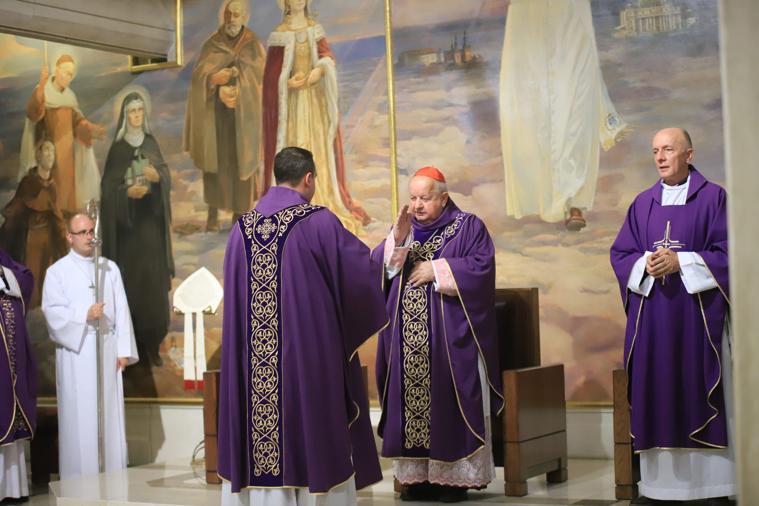 A Catholic cardinal in purple robes makes the sign of the cross in front of another member of the Catholic clergy, who is wearing similar purple robes. There is a mural of various Catholic figures and saints in the background.