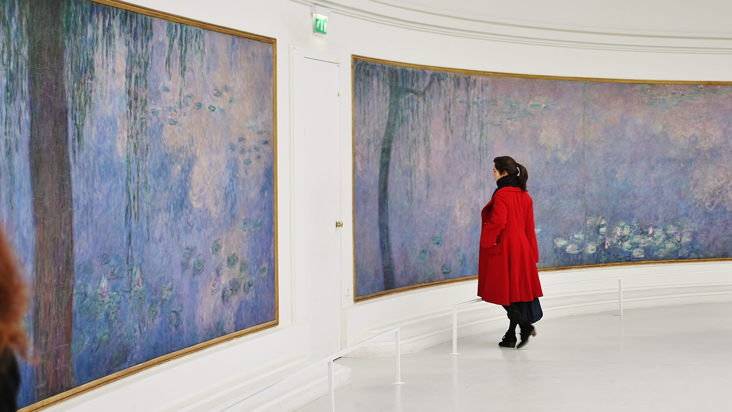 A dark-haired woman in a vibrant red coat admires a set of impressionistic water lillies paintaings by Claude Monet. The paintings feature weeping tress and water lillies on violet and pink water.