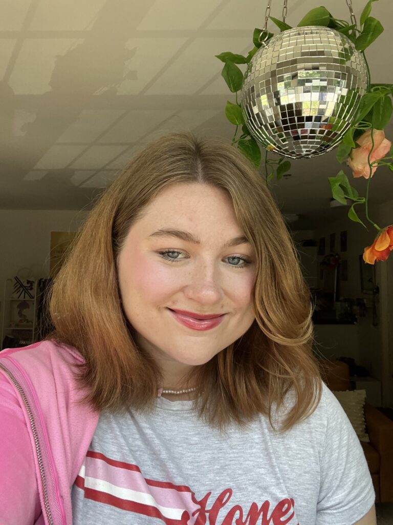 Cropped photo of Jaylin, a white woman with shoulder-length brown-blond hair, smiling at the camera. There is a disco ball with a fake plant draped over it in the background.